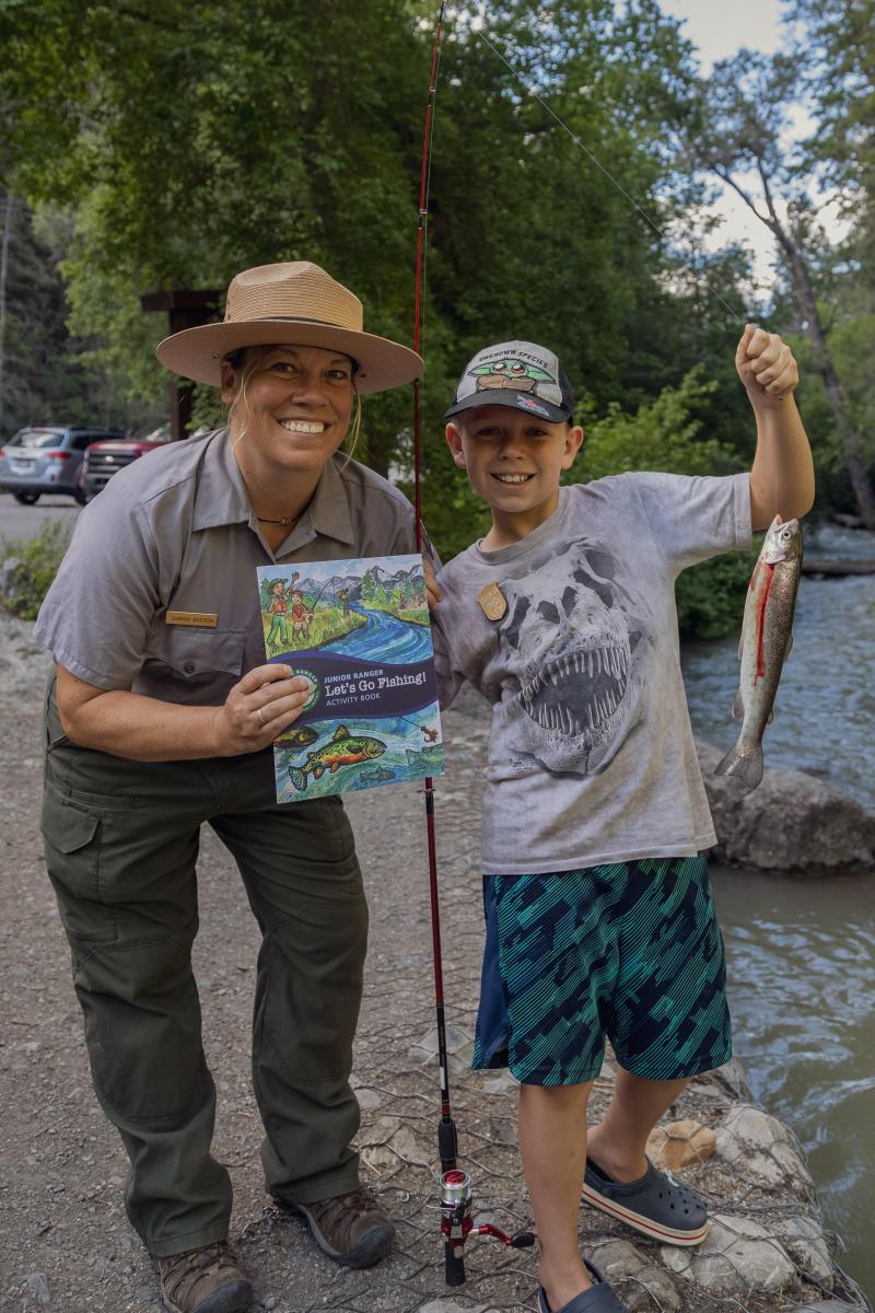 Ranger posing by child with a fish, fishing pole, and Junior Ranger book