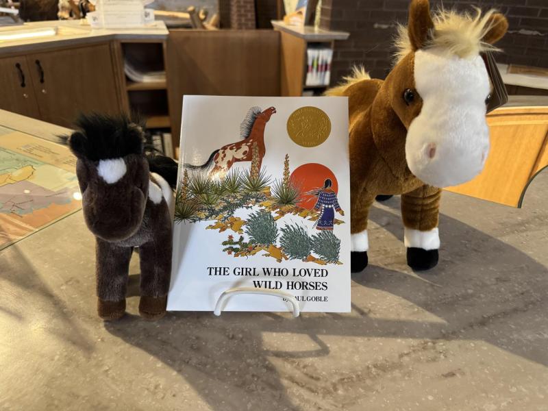 Two stuffed animal horses stand on either side of the book "The Girl Who Loved Horses" by Paul Goble