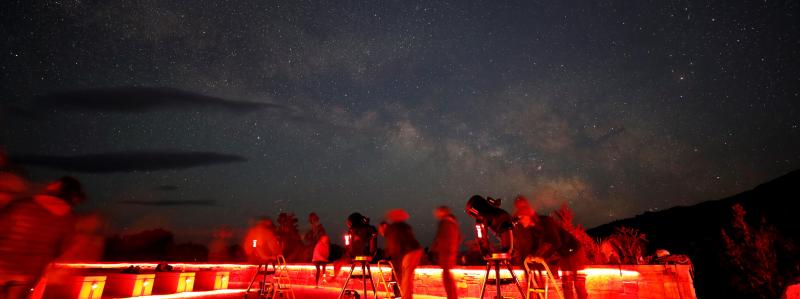 Three telescopes with shadowy figures due to long exposure stand in front of a colorful Milky Way.