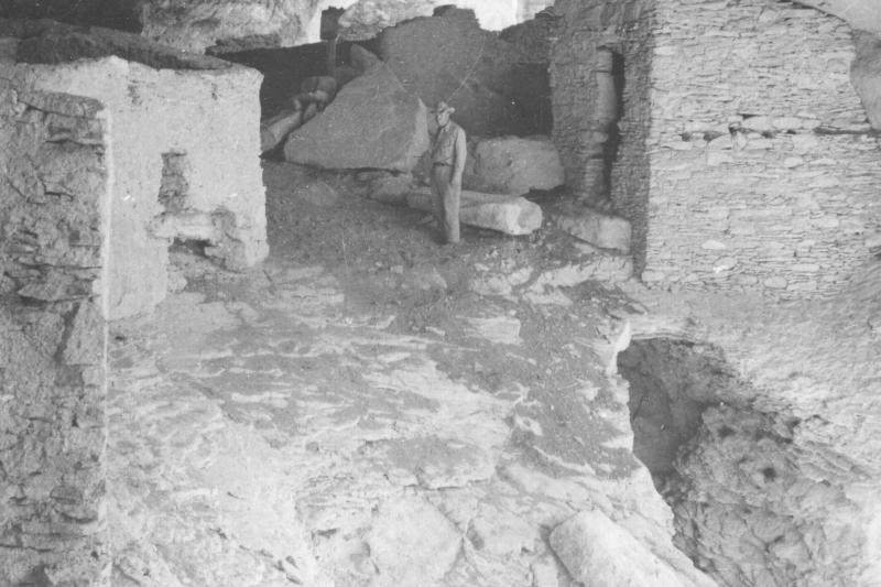 Black and white photo of stone houses in a cliff with a man wearing a ranger uniform.