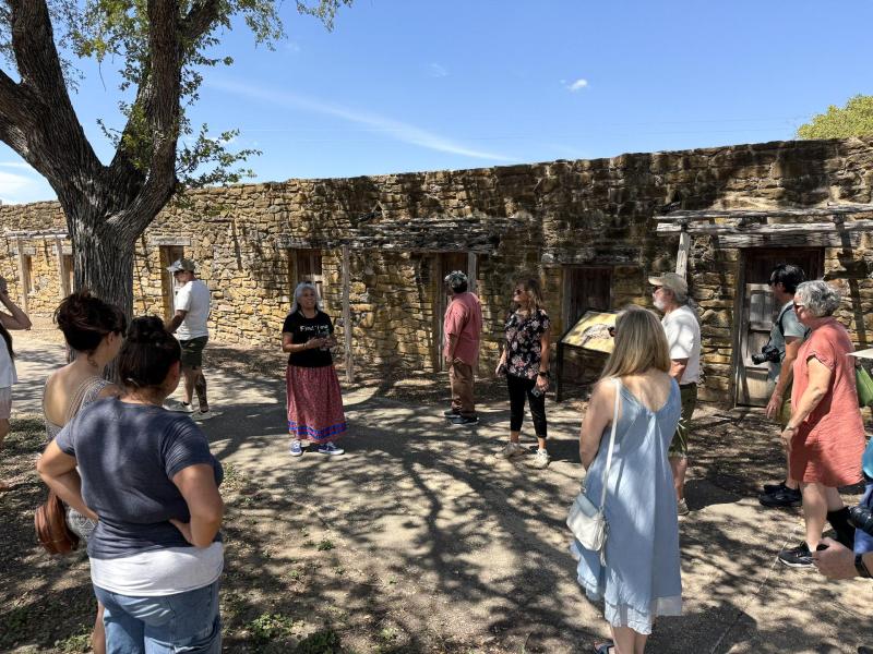 Group of people gathered outdoors near a historic stone building under a clear blue sky with tree sh