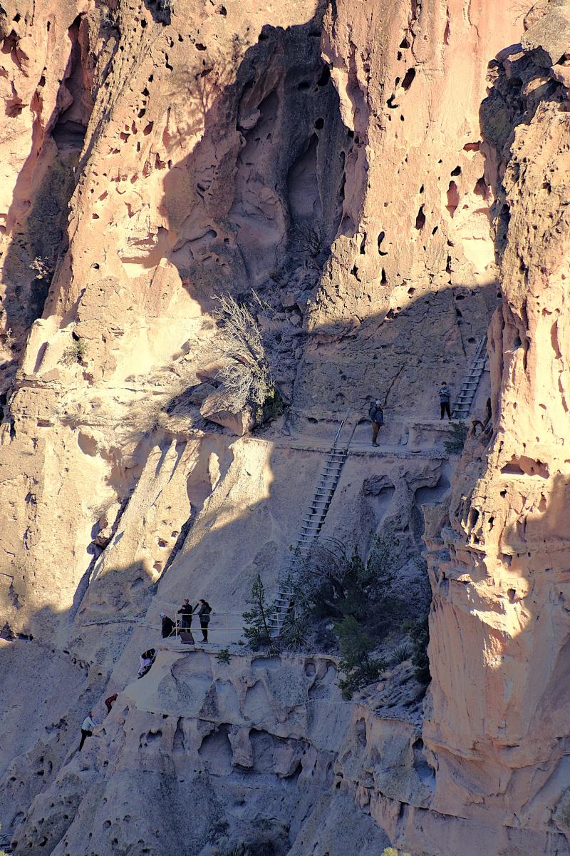 People stand beneath a series of tall, wooden ladders climbing a cliff to an alcove.