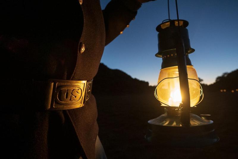 A person in reenactor clothing, including a gold belt buckle that says "US" holds a lantern at dusk