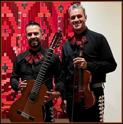 Two men in black mariachi outfits stand in front of a red patterned backdrop.