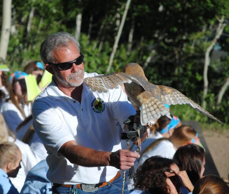 A man in a white shirt and sunglasses holds an owl perched on his outstretched arm.