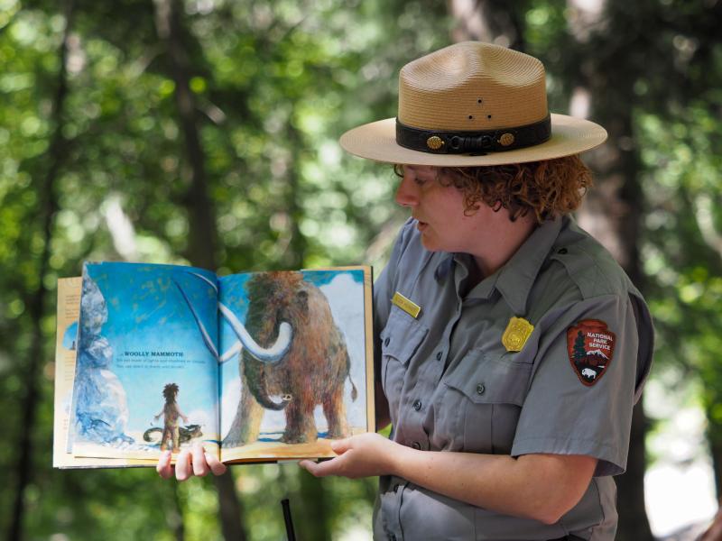 A park ranger holds a book open to her side while reading to others in a forested setting.
