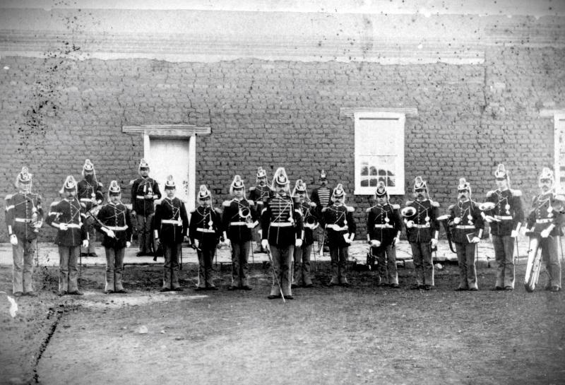 An 1800s image of a uniformed Army infantry band standing in front of an adobe wall.