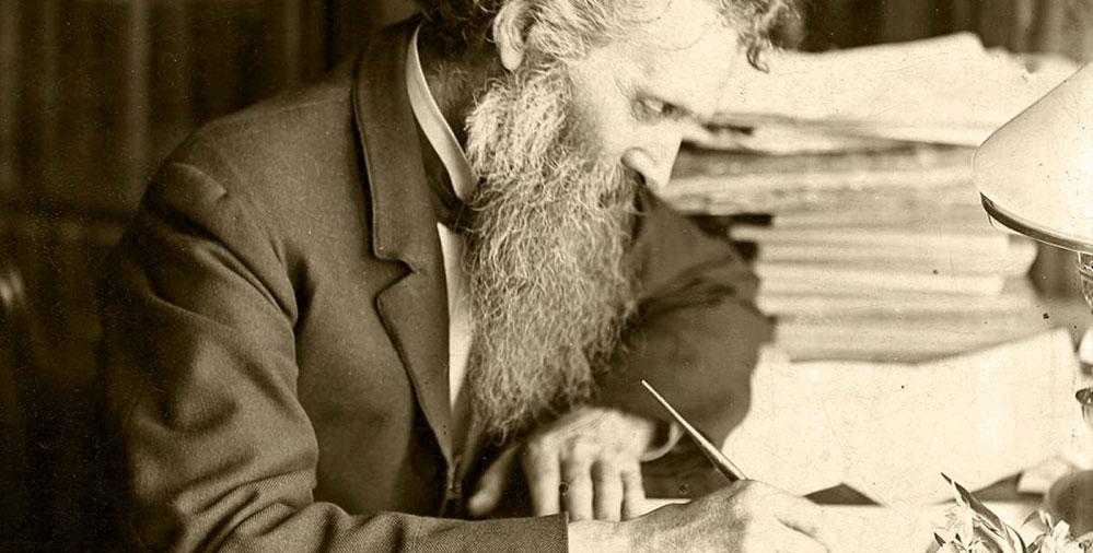John Muir sits at his desk and writes. The desk is displayed in the 