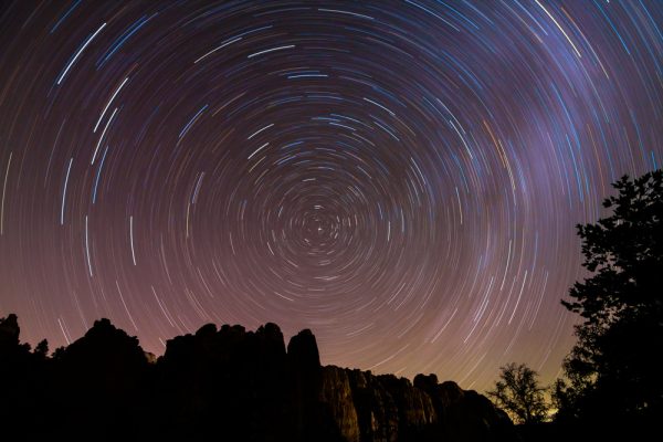 Star trails around the center of the image with silhouetted trees and rocks in the foreground.