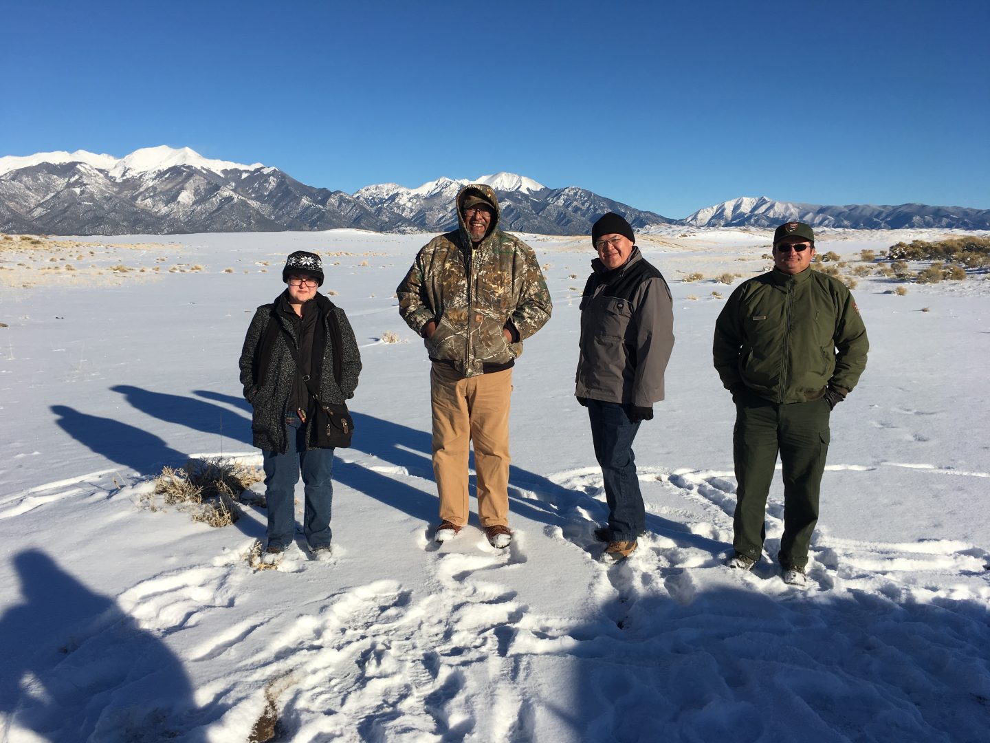 Tesuque Pueblo members stand with NPS staff in a remote desert area of the park in winter, with snow on the ground. In the distant background are snowy dunes and snow-capped mountains.